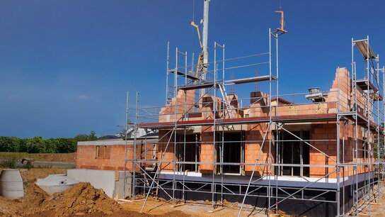 Baustelle mit zwei unvollendeten Häusern, umgeben von Baugerüst und Erdarbeiten unter blauem Himmel.