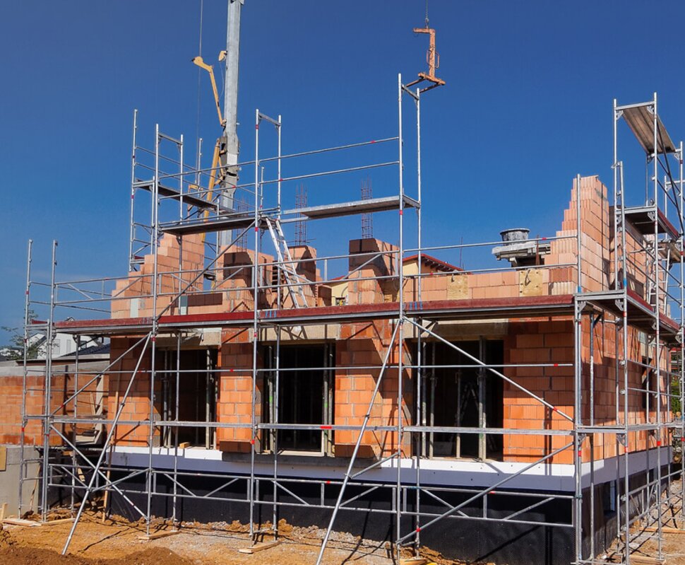 Baustelle mit zwei unvollendeten Häusern, umgeben von Baugerüst und Erdarbeiten unter blauem Himmel.