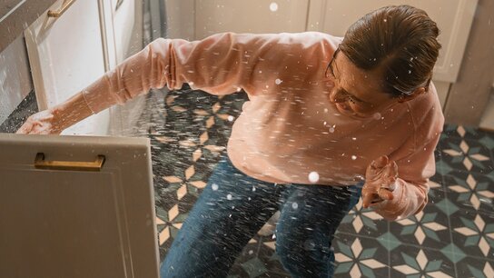 Person kniet vor einem offenen Schrank, während Wasser spritzt und den Küchenboden durchnässt.