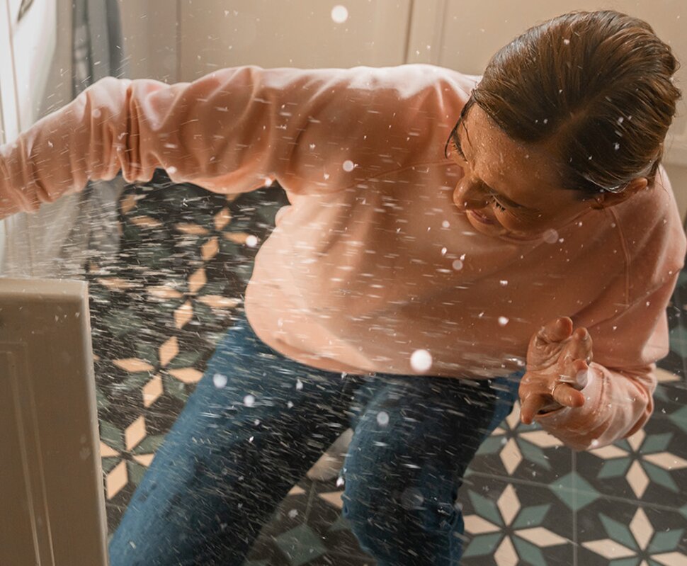 Person kniet vor einem offenen Schrank, während Wasser spritzt und den Küchenboden durchnässt.