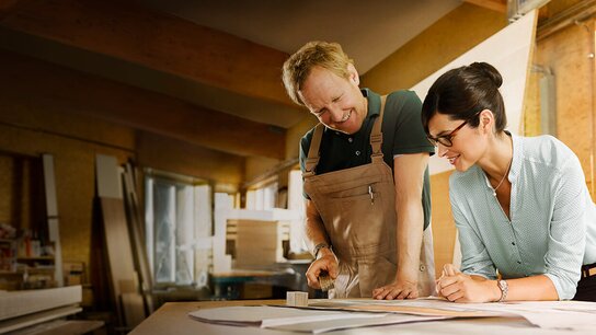 Ein Handwerker und eine Frau betrachten Pläne auf einem Tisch in einer Werkstatt, umgeben von Materialien und Werkzeugen.