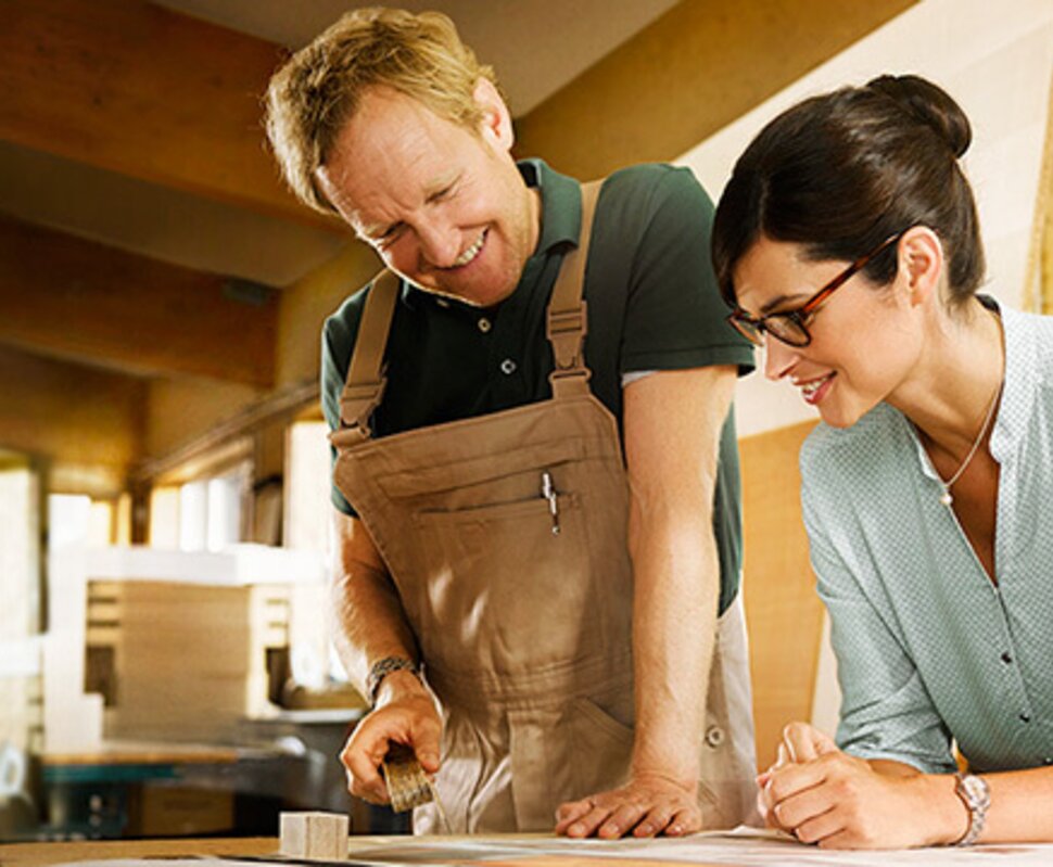 Ein Handwerker und eine Frau besprechen Pläne an einem Tisch in einer hellen Werkstatt. Beide wirken engagiert und freundlich.