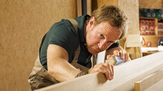 Handwerker arbeitet konzentriert an einem Holzstück und verwendet dabei ein Werkzeug zur Bearbeitung der Oberfläche.