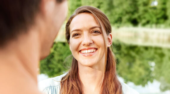 Lächelnde Frau betrachtet einen Mann, im Hintergrund spiegelt sich ein ruhiger See und üppige grüne Bäume.