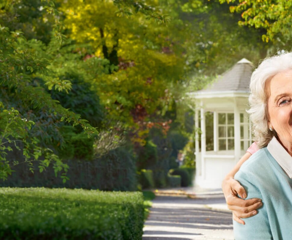 Zwei lächelnde Frauen posieren im Garten für ein Selfie, umgeben von grünen Bäumen und einem weißen Pavillon.