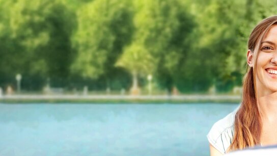Lächelnde Frau im Vordergrund, steht am Wasser und blickt freundlich zu einer Person außerhalb des Bildes. Grünes Ufer im Hintergrund.