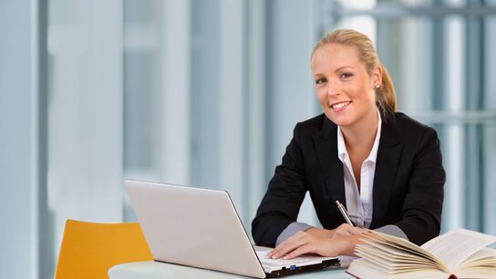 Junge Frau in Anzug sitzt an einem Tisch mit Laptop und Buch, lächelt in einem modernen Büro.