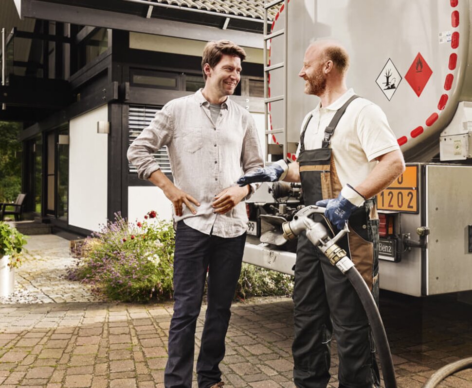 Zwei Männer im Gespräch neben einem Lieferwagen auf einem gepflasterten Außenbereich eines modernen Hauses mit Garten.