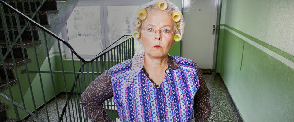 Ältere Frau mit Haarwicklern und schockiertem Gesichtsausdruck steht in einer grünen Treppe.