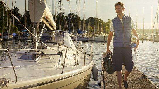 Mann mit einem Rucksack und einer Jacke steht an einem Yachthafen neben einem Segelboot. Sonne scheint am Abend.