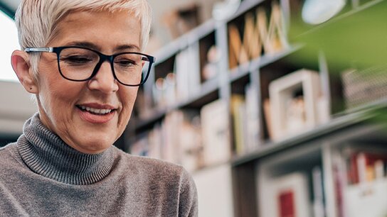 Eine ältere Frau mit kurzen, grauen Haaren und Brille lächelt, während sie auf einem Laptop arbeitet. Pflanzen im Vordergrund.