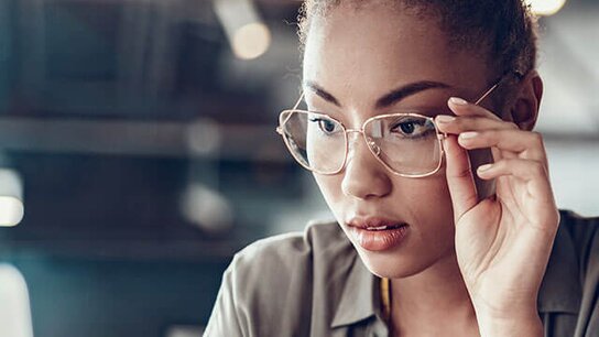Eine junge Frau mit lockigem Haar trägt Brillen und schaut konzentriert auf einen Laptop, daneben eine Kaffeetasse.