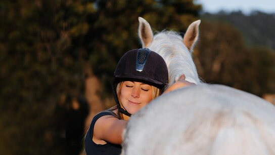 Ein Mädchen in einem Reithelm umarmt ein weißes Pferd, lächelnd und zufrieden in einer natürlichen Umgebung.