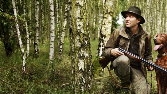 Mann im Wald, bekleidet mit Jagdausrüstung, steht auf einem umgefallenen Baumstamm und hält ein Gewehr in der Hand.