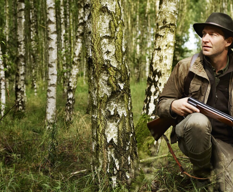 Mann im Wald, bekleidet mit Jagdausrüstung, steht auf einem umgefallenen Baumstamm und hält ein Gewehr in der Hand.