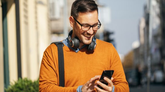 Mann mit Brille und Kopfhörern in orangefarbenem Pullover, der lächelnd auf sein Smartphone schaut.