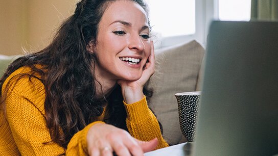 Lachende Frau in orangem Pullover, die entspannt vor einem Laptop liegt und den Bildschirm aufmerksam betrachtet.