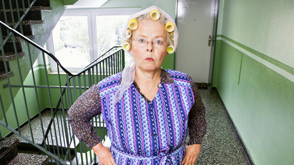 Ältere Frau mit Lockenwicklern, in bunter Schürze, steht in einem Treppenhaus mit grünen Wänden und schaut ernst.