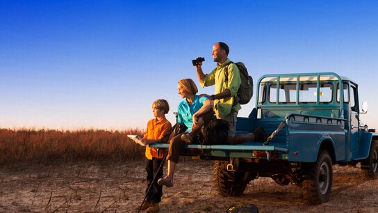 Familie mit einem Kind sitzt auf dem Heck eines blauen Geländewagens in einer weiten, trockenen Landschaft bei Sonnenuntergang.