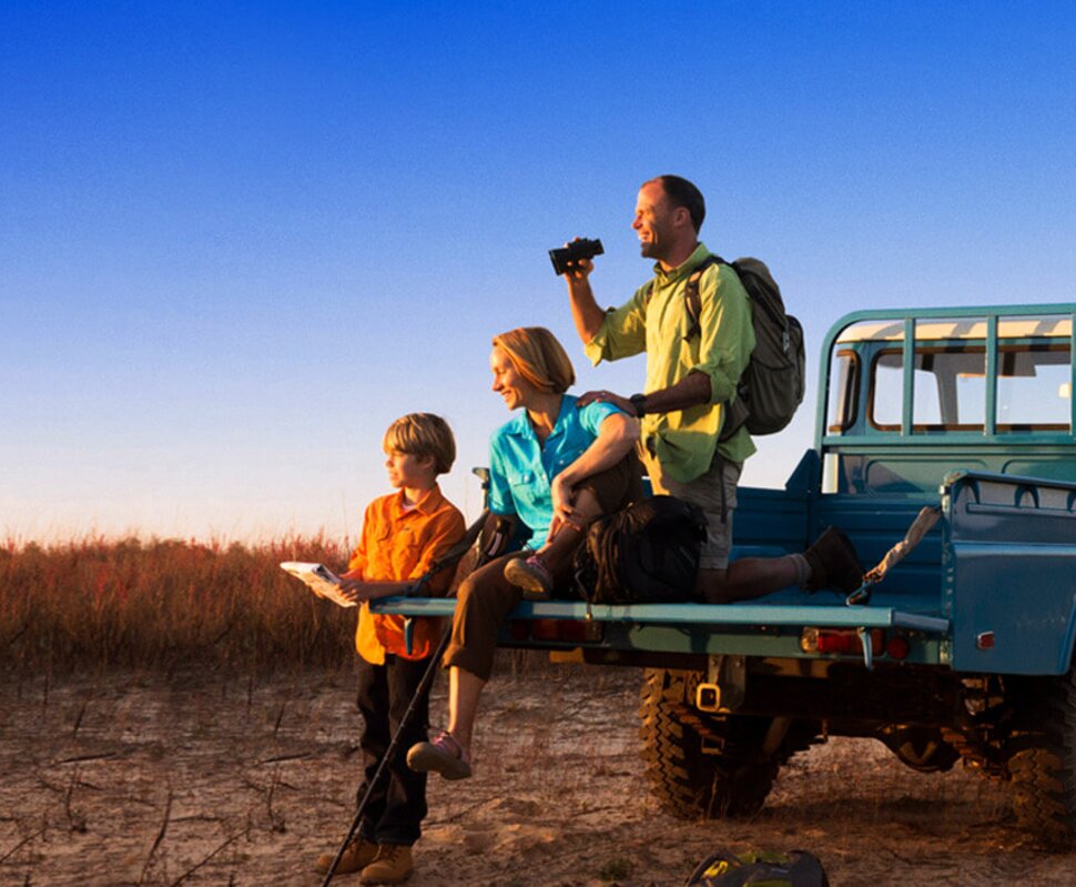 Familie mit einem Kind sitzt auf dem Heck eines blauen Geländewagens in einer weiten, trockenen Landschaft bei Sonnenuntergang.