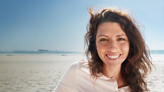 Lächelnde Frau mit lockigem Haar vor einem sonnigen Strand, sanfte Wellen und blauer Himmel im Hintergrund.