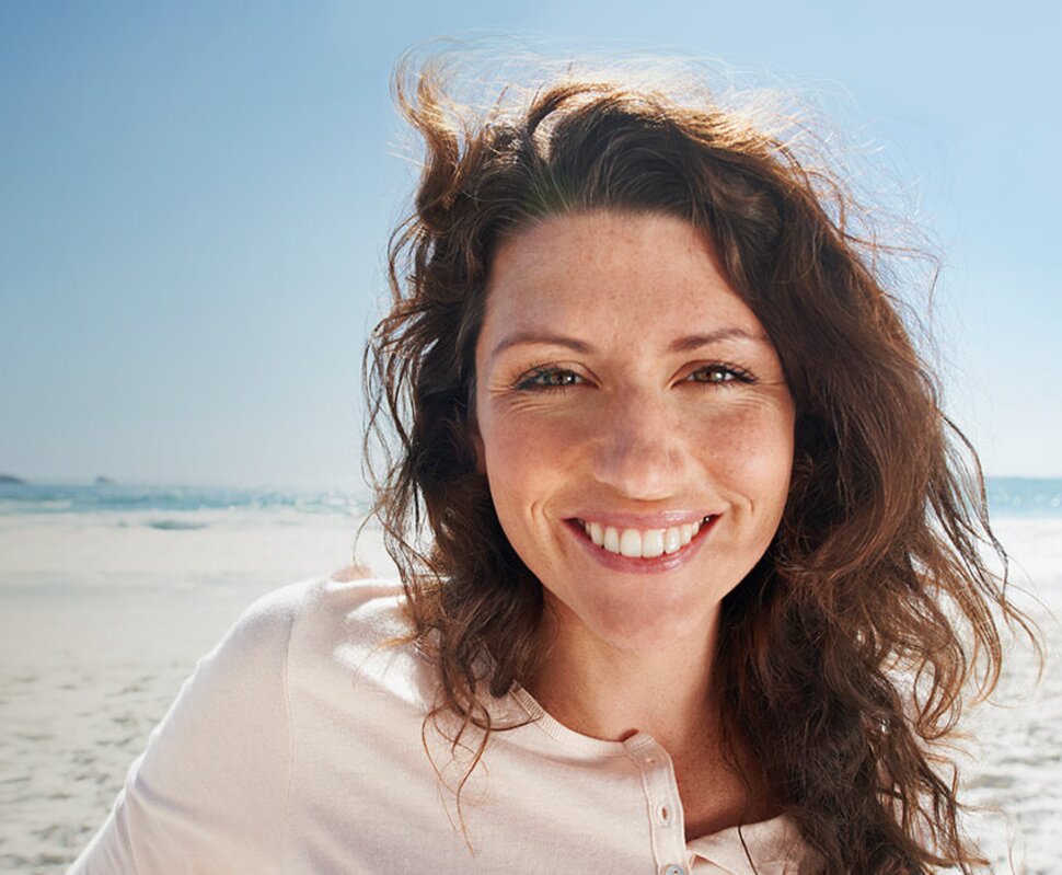 Lächelnde Frau mit lockigem Haar vor einem sonnigen Strand, sanfte Wellen und blauer Himmel im Hintergrund.