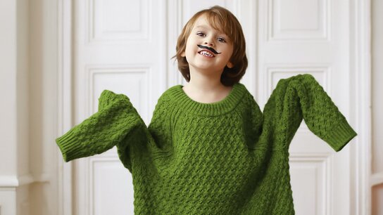 Ein fröhliches Kind in einem oversized grünen Pullover mit einer Spielzeugmustache auf dem Gesicht, das lacht.