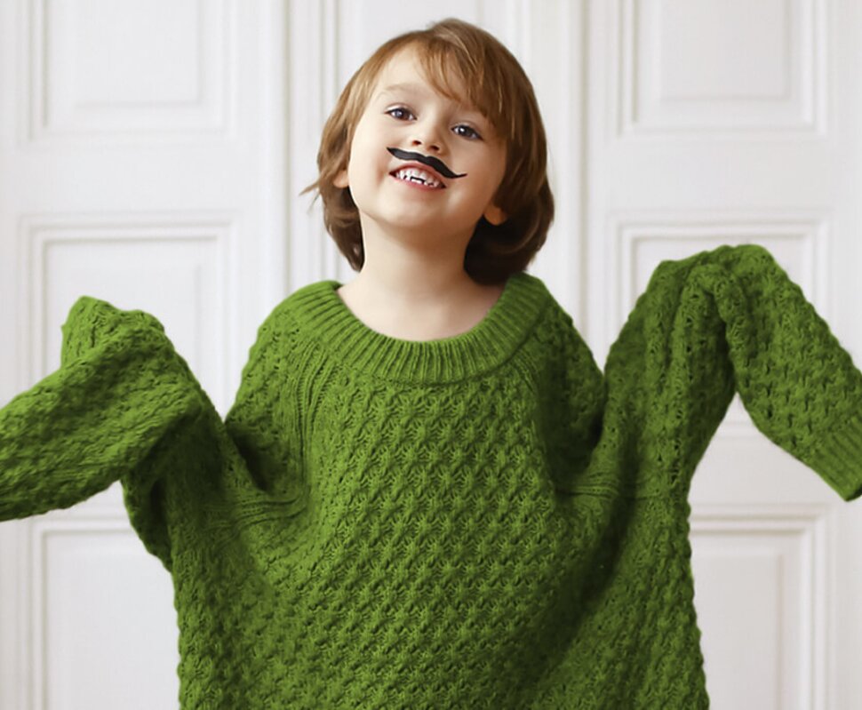 Ein fröhliches Kind in einem oversized grünen Pullover mit einer Spielzeugmustache auf dem Gesicht, das lacht.