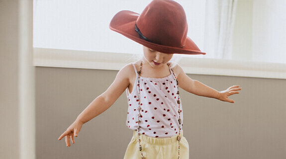 Kleines Kind mit rotem Hut und gepunktetem Top, das fröhlich in einem hellen Raum steht und mit den Armen spielt.