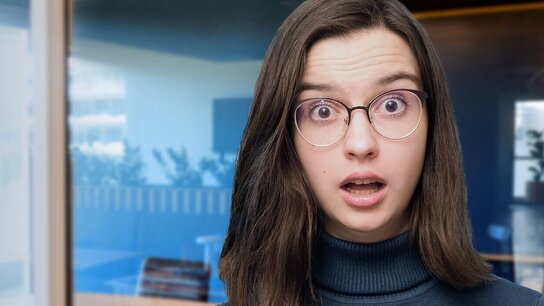 Junge Frau mit Brille und überraschter Miene steht in einem modernen Büroraum.