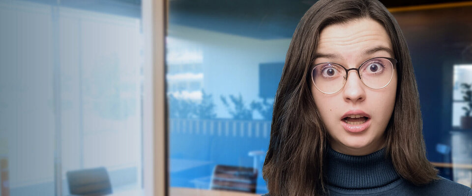Junge Frau mit Brille und überraschter Miene steht in einem modernen Büroraum.