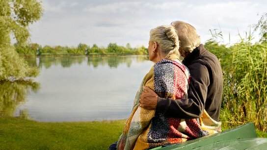 Ein älteres Paar sitzt auf einer Bank am Wasser, umarmt und blickt gemeinsam über die ruhige Oberfläche des Sees.