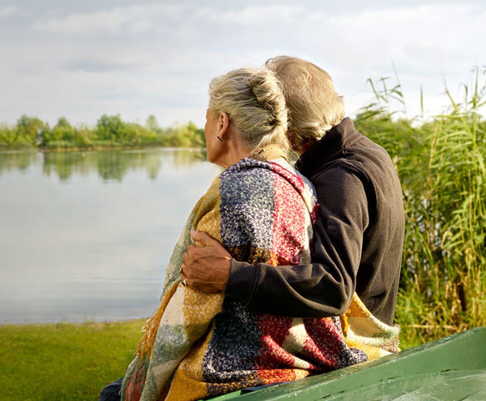 Ein älteres Paar sitzt auf einer Bank am Wasser, umarmt und blickt gemeinsam über die ruhige Oberfläche des Sees.