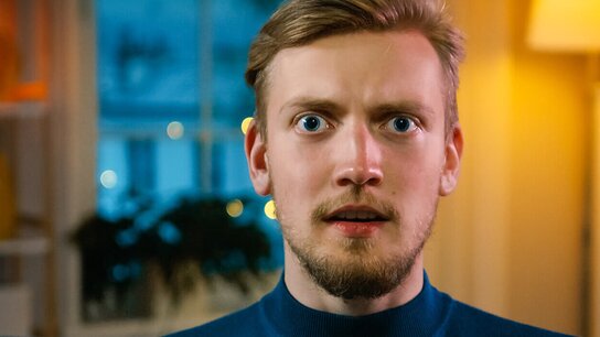 Junger Mann mit shocked Ausdruck, Blick auf die Kamera gerichtet, im Hintergrund unscharfe, warme Lichter und Fenster.