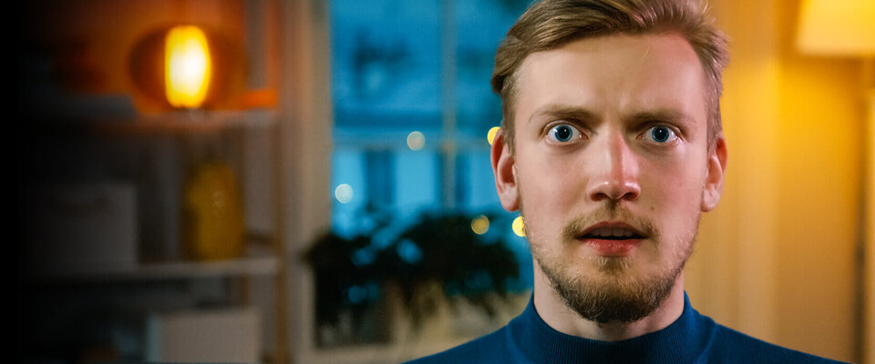 Junger Mann mit shocked Ausdruck, Blick auf die Kamera gerichtet, im Hintergrund unscharfe, warme Lichter und Fenster.