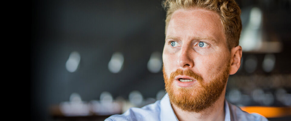 Ein Mann mit blondem Haar und rotbraunem Bart schaut überrascht. Im Hintergrund unscharfe, helle Lichtpunkte.