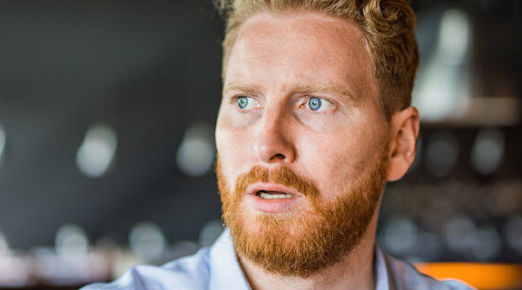 Mann mit blonden lockigen Haaren und Bart, schaut nachdenklich in die Ferne, Hintergrund unscharf.